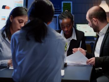 Diverse business entrepreneurs sitting at conference table working at management solution presentation in office meeting room late at night. Multi ethnic teamwork brainstorming ideas